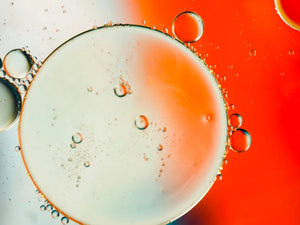 Illustration macro de bulles et phase aqueuse orangée, associée à l’acide lactique (AHA), un exfoliant doux utilisé en cosmétique pour lisser le grain de peau et améliorer l’éclat.