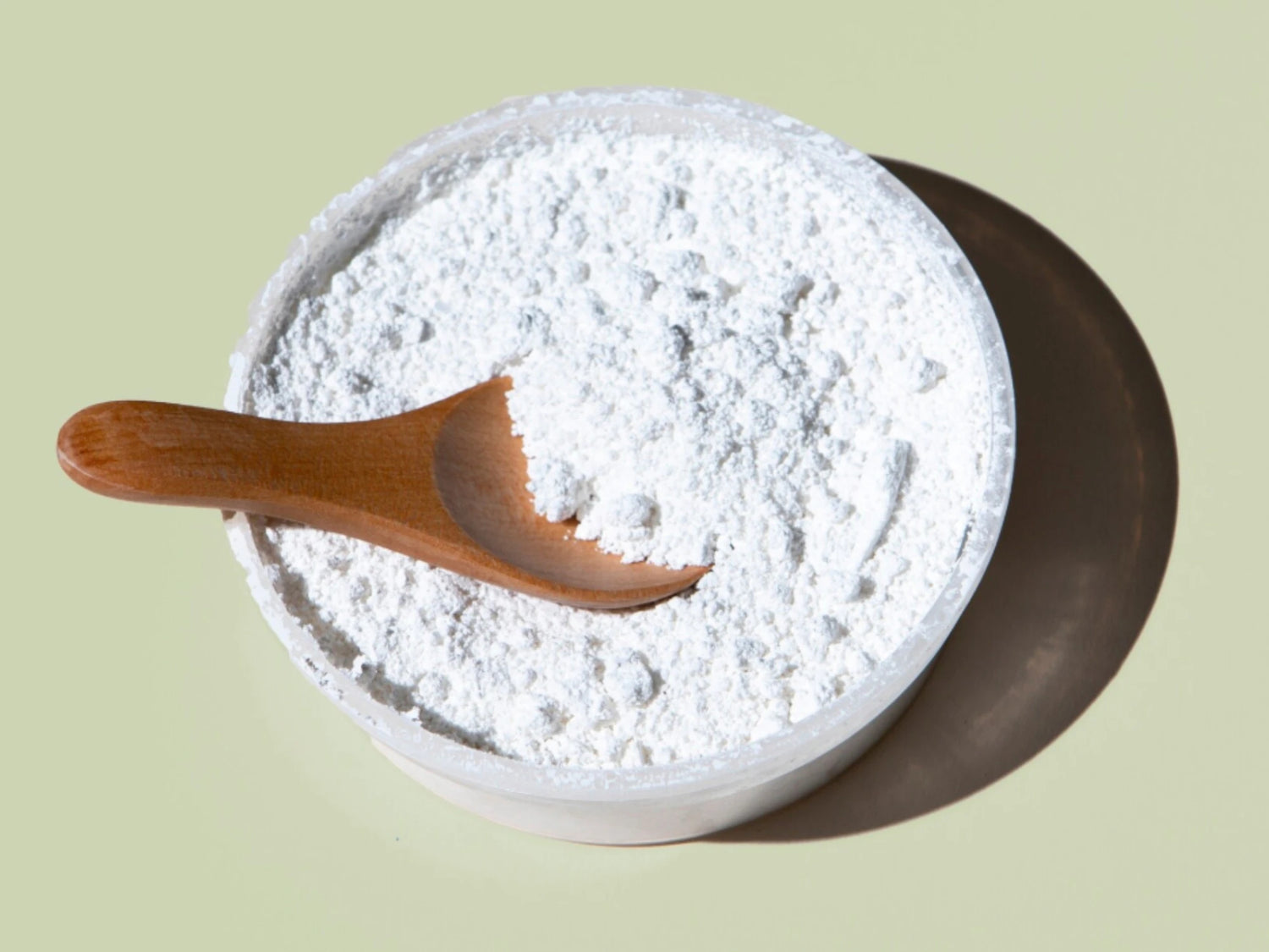 Bol de poudre blanche avec cuillère en bois, illustration de l’Ascorbyl Glucoside (dérivé de vitamine C stabilisée) utilisé en cosmétique pour soutenir l’apparence d’un teint plus lumineux et homogène.