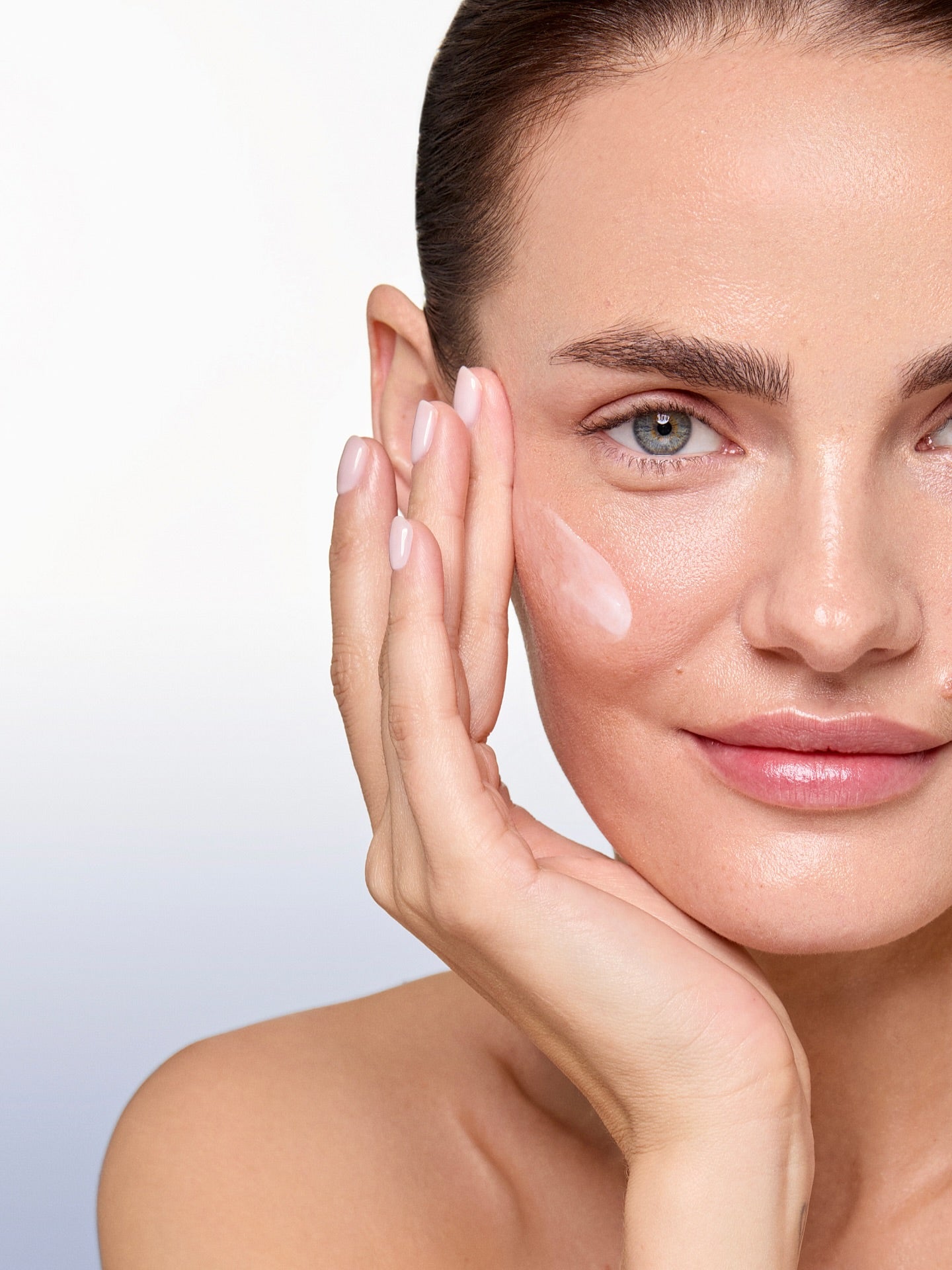 Femme appliquant un soin visage sur la joue, texture crème, routine hydratante sur fond blanc.